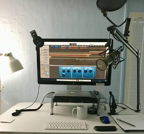 Una mesa de trabajo con un ordenador, micrófono de condensador, auriculares y libreta y boli
